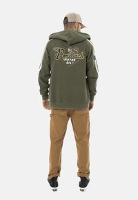Sweat à capuche vert avec capuche et motif graphique au dos, associé à un pantalon cargo beige et des baskets noires. Tissu doux, design sportif.