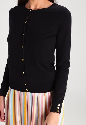 Mujer con un cárdigan negro abotonado con botones dorados y una falda con rayas verticales multicolores.