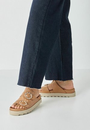 Personne portant un jean bleu foncé et des sandales marron clair avec des boucles ornées de bijoux, debout sur un sol clair.