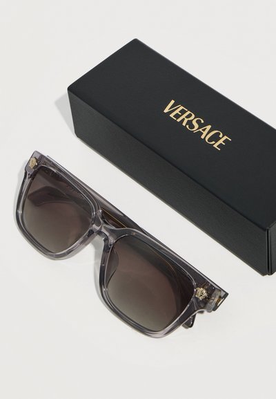 Γκρι αποχρώσεων τετράγωνα γυαλιά ηλίου Versace με το έμβλημα της Μέδουσας κοντά στις μεντεσέδες, τοποθετημένα δίπλα σε μαύρη θήκη με χρυσή σήμανση Versace πάνω σε λευκή επιφάνεια.