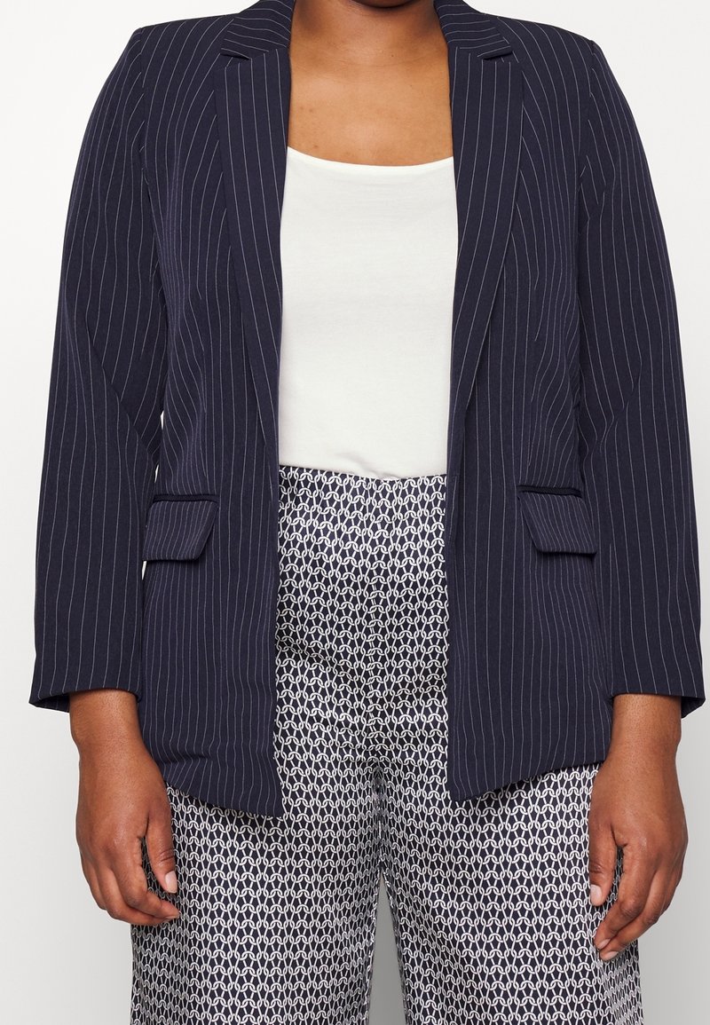Personne portant un blazer bleu marine à fines rayures sur un haut blanc et un pantalon noir et blanc à motifs, debout, les bras relâchés le long du corps.
