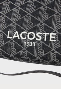 Czarna torba z wzorzystego materiału z białym logo "LACOSTE" oraz detalem "1933". Posiada gładką teksturę i cienki pasek. Projekt zawiera powtarzające się motywy.