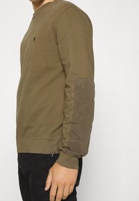 Olivgrön sweatshirt med en mjuk textur, med en enfärgad kropp och en kontrasterande tygpatch på ärmarna. Ribbad mudd och nederkant.