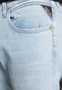 Ljust blå denimjeans med en strukturerad yta, som har en bakficka med en diskret logotagg och en guldknytt