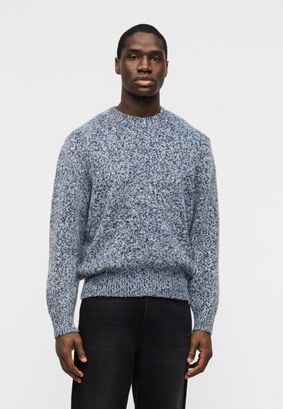 Homme portant un pull en maille bleu et blanc chiné et un pantalon noir, debout devant un fond gris clair uni.