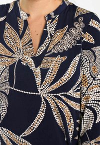 Granatowa bluzka z kwiatowym wzorem w bieli i złocie, z dekoltem w serek i gładkim, lekkim materiałem.