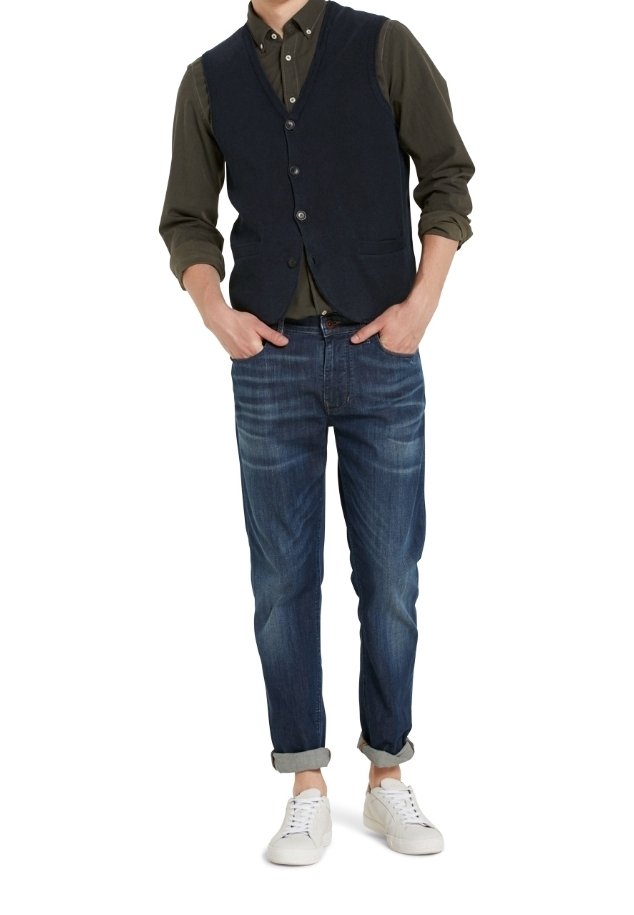 Granatowa kamizelka z guzikami na ciemnozielonej koszuli, zestawiona z jasnoniebieskimi dżinsami i białymi sneakersami. Casualowy styl z podwiniętymi mankietami.