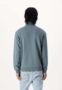 Persona con capelli scuri che indossa una felpa a maniche lunghe color teal e jeans azzurri chiari, girata di spalle contro uno sfondo bianco semplice.