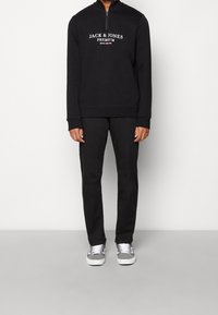 Czarna bluza z zamkiem z białym logo, w połączeniu z czarnymi spodniami i szarymi sneakersami. Gładki materiał, luźny krój, minimalistyczny design.