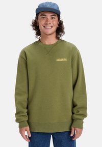 Olivovo zelená mikina s voľným strihom, rebrovanými manžetami a žltým logom Quiksilver. Nosená s modrými denimovými džínami a svetlomodrou čiapkou.