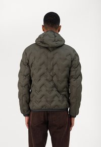 Giacca a vento verde oliva con cappuccio, caratterizzata da una trama trapuntata e rifiniture nere ai polsini e all'orlo. Indossata sopra pantaloni marroni.