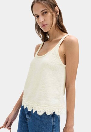 Donna che indossa un top senza maniche in maglia cremosa con orlo in pizzo smerlato, abbinato a jeans blu, che tiene una borsa marrone con tracolla su uno sfondo uniforme.