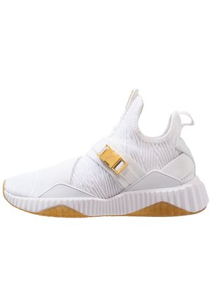 Basket de sport montante blanche avec tige en tissu texturé, sangle avec boucle dorée et semelle brune mouchetée.