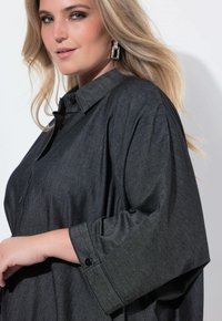Blonde Frau mit offenen Haaren trägt geometrische silberne Ohrringe und ein dunkelgraues Hemd mit breiten Ärmeln und posiert vor einem weißen Hintergrund.