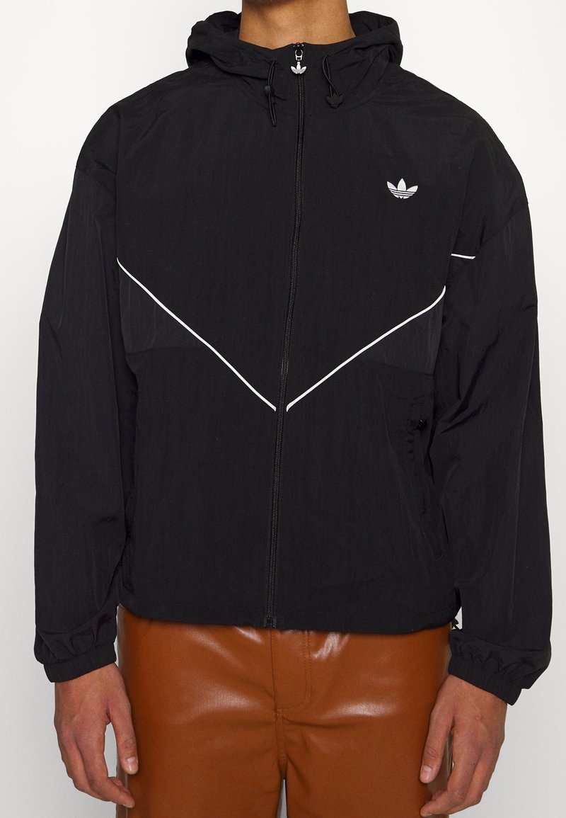 Veste légère noire avec capuche, arborant des rayures diagonales blanches et un logo brodé sur le côté gauche de la poitrine. Poignets et ourlet élastiques.