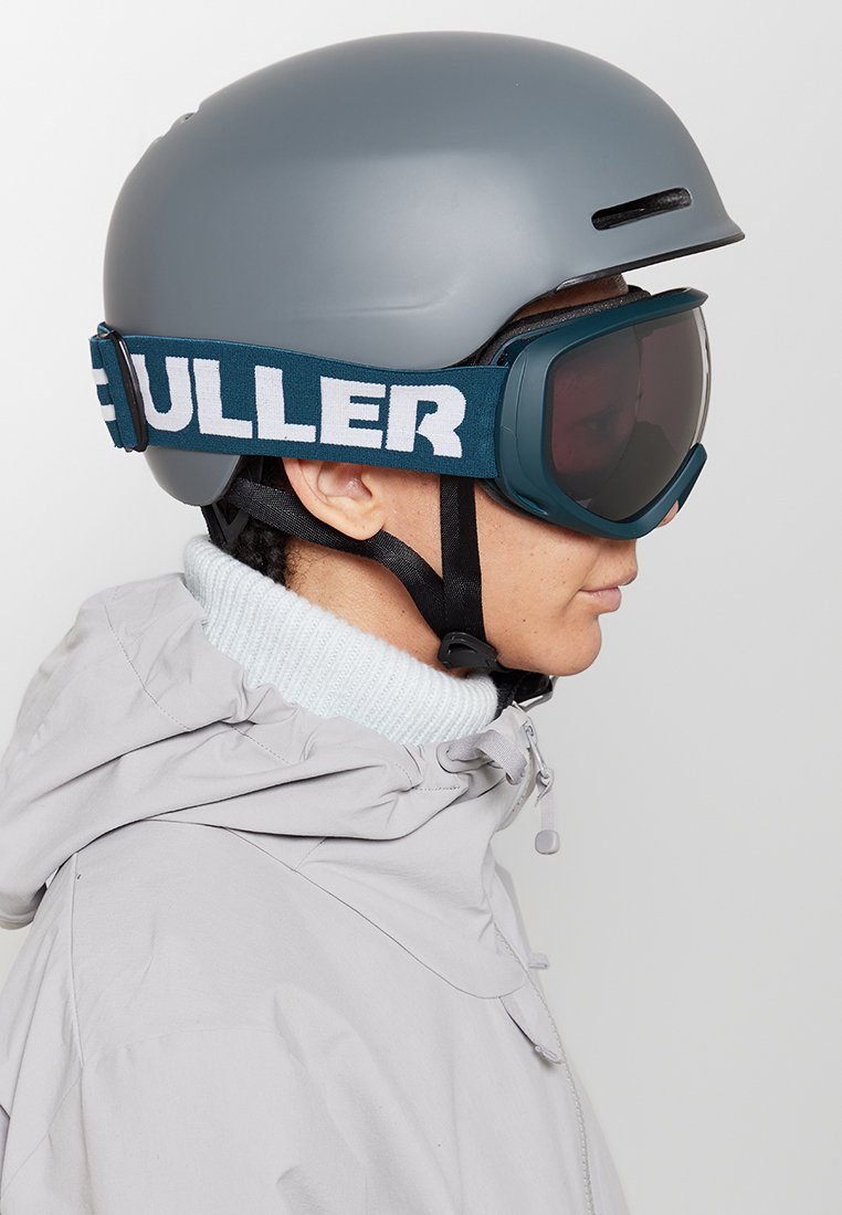 Grijze skihelm met een matte afwerking, gecombineerd met donkerblauwe skibril met een brede band met witte tekst. De drager heeft een lichtjack aan.