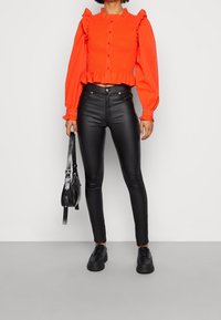 Blusa rossa arricciata con maniche a volant, abbinata a jeans skinny in pelle nera e scarpe stringate nere. Include una borsa a tracolla.