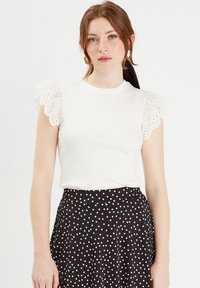 Top blanc à côtes avec des manches à volants en dentelle, associé à une jupe à pois noirs. Le tissu a une texture lisse et un design ajusté.