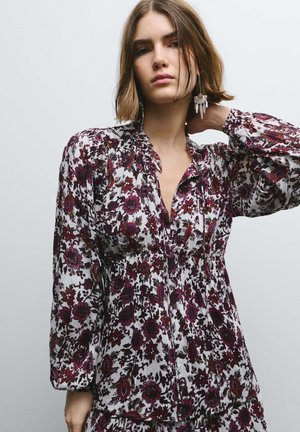 Lange mouwen jurk van witte stof met een paars en rood bloemenpatroon, met een geplooide taille en een gerimpelde halslijn.