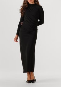 Robe noire à manches longues avec un design drapé, pr�ésentant des fronces sur le côté et une silhouette élégante et ajustée. Portée avec des escarpins à bout pointu.