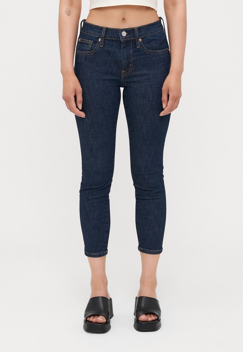 Jeans skinny de mezclilla azul oscuro con una cintura alta, diseño de cinco bolsillos y costuras sutiles en naranja. Combinados con sandalias negras de plataforma.