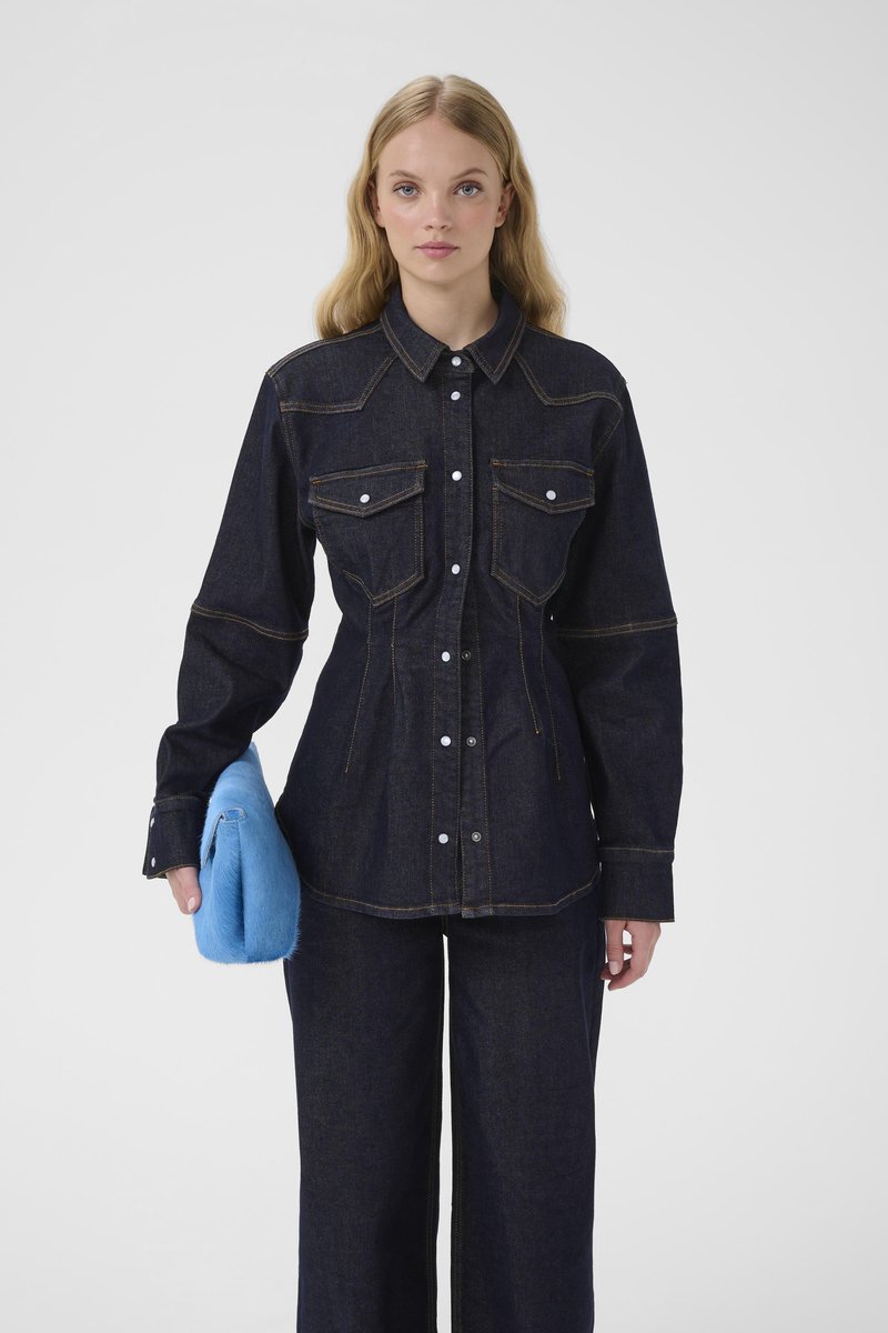 Mørk denimskjorte med to brystlommer, kontrasterende gule syninger og knaplukning. Kombineret med viddebenede denimbukser og en blå pelsclutch.