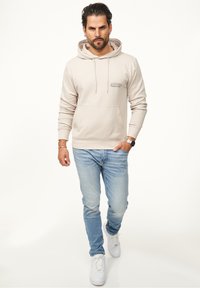 Beige hooded sweatshirt with a front kangaroo pocket and embroidered logo. Paired with light blue jeans and white sneakers.