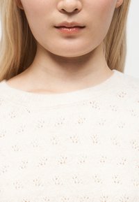 Gros plan sur le bas du visage et le cou d'une personne portant un pull en maille blanc avec un motif délicat.