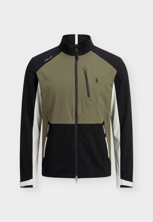 Giovane uomo in piedi che indossa una giacca con zip verde, nera e bianca, pantaloni neri e scarpe da ginnastica bianche su uno sfondo grigio chiaro uniforme.