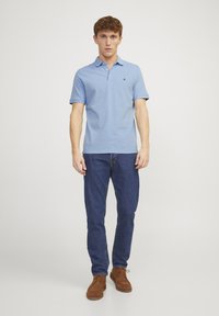 Blaues Poloshirt mit Kragen und Knopfleiste mit drei Knöpfen, kombiniert mit dunkelblauen Jeans und braunen Wildlederschuhen. Schlichte Gestaltung, keine Muster.