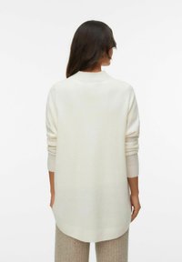 Sweater surdimensionné blanc avec un col haut, des poignets côtelés et des épaules tombantes, associé à un pantalon en tricot beige texturé.