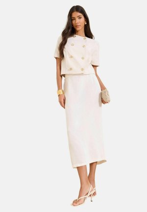 Woman wearing a cream double-breasted short-sleeve jacket, matching midi skirt, gold cuff bracelet, woven clutch, and nude strappy heels.