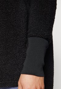 Black fleece jacket fabric with textured surface. Cuffs in a smooth black knit material. Close-up view emphasizes contrasting textures.