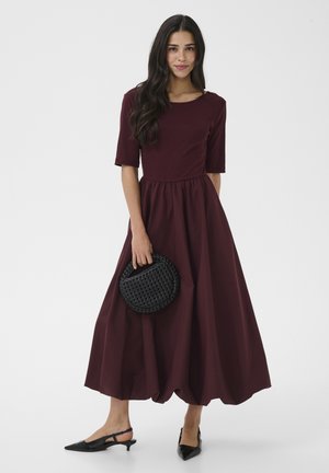 Femeie purtând o rochie midi bordo cu mâneci până la cot, ținând un clutch circular negru țesut și purtând pantofi negri cu barete pe călcâi.