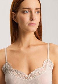 Lace-trimmed light beige camisole with thin straps, featuring a ribbed texture and a scalloped edge at the neckline.