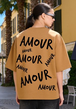 Femme aux cheveux foncés attachés en queue de cheval et lunettes de soleil porte une chemise marron imprimée plusieurs fois avec le mot "AMOUR" en police noire.