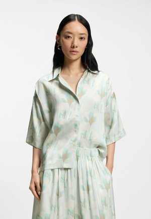 Frau mit langen schwarzen Haaren, die eine lockere, hellgrüne blumige Bluse mit Knöpfen und einen passenden Rock trägt, steht mit einer Hand in der Tasche.