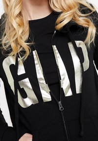 Sudadera con cremallera negra y gran lettering metálico dorado "GAP", con capucha ajustable y una textura de tela suave.
