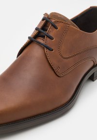 Zapato de cuero marrón con un acabado liso, que presenta una puntera moldeada, costuras decorativas y cordones en color marino. Suela en escala de grises con ligera elevación.