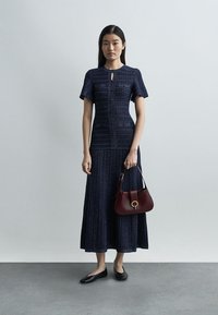 Γυναίκα που φορεί ένα σκούρο μπλε υφασμάτινο φόρεμα, κρατώντας μια μπορντό τσάντα, στέκεται σε γκρι πάτωμα μπροστά από έναν ανοιχτό γκρι τοίχο.