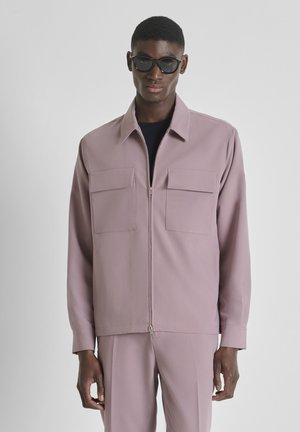 Man draagt mauve zip-up jack met twee borstzakken en bijpassende broek, geaccentueerd met zwarte zonnebril, staand tegen een effen achtergrond.