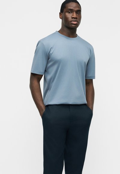 Νεαρός άνδρας που φοράει ανοιχτό μπλε κοντομάνικο μπλουζάκι και σκούρο παντελόνι, στέκεται με τα χέρια στις τσέπες μπροστά σε ένα απλό φόντο.