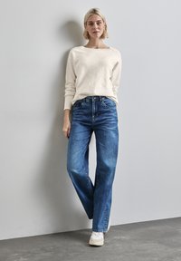 Maglione chiaro con scollatura a costine, abbinato a jeans blu larghi e sneakers bianche. I jeans presentano un classico stile a cinque tasche.