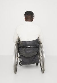 Černý invalidní vozík se stříbrnými koly, s černým sedákem a opěradlem. Osoba v bílé texturované mikině sedící zády.