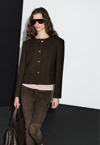 Vrouw met lang golvend haar, gekleed in een donkerbruine knopenjas, suede broek, grote zonnebril en een bijpassende bruine leren tas.