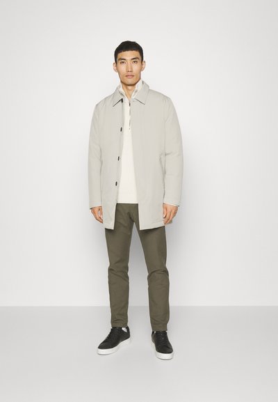 Abrigo gris claro con un cuello, botones negros y cierre delantero, llevado sobre un suéter crema y pantalones oliva, combinado con zapatillas negras.
