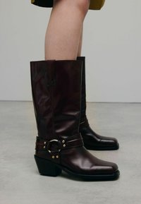 Bottes en cuir marron arrivant au genou avec un bout carrée, deux sangles et un accent en anneau métallique. Fini texturé avec un léger talon.