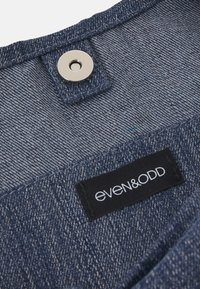 Gros plan sur un tissu denim bleu avec un bouton-pression en métal et une étiquette en tissu noire portant l’inscription « even&odd » cousue dessus.