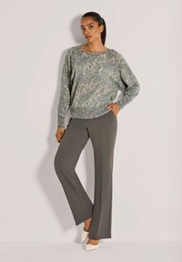 Vrouw die staat in een gebreide lange mouw trui met patroon, grijze wijde broek en witte puntige hakken tegen een effen achtergrond.