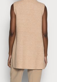 Gilet sans manches beige en tricot avec une texture côtelée, doté d'un col montant et d'une coupe décontractée, descendant en dessous de la taille.